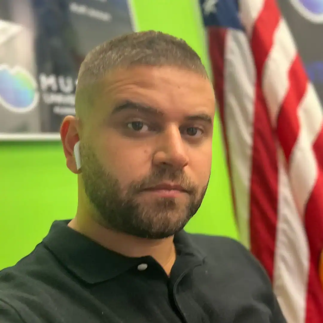 Homem com máscara de proteção facial e fone de ouvido, em ambiente com fundo verde e bandeira dos EU.